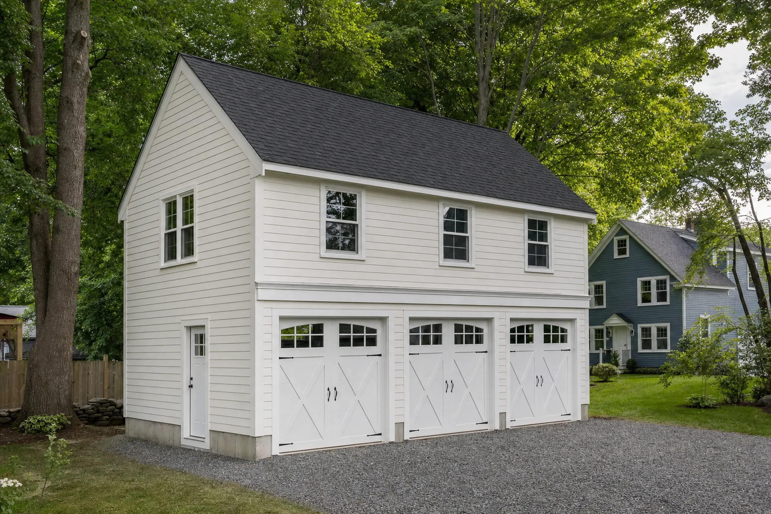White two-story garage with three doors and black roof in a green yard with trees.