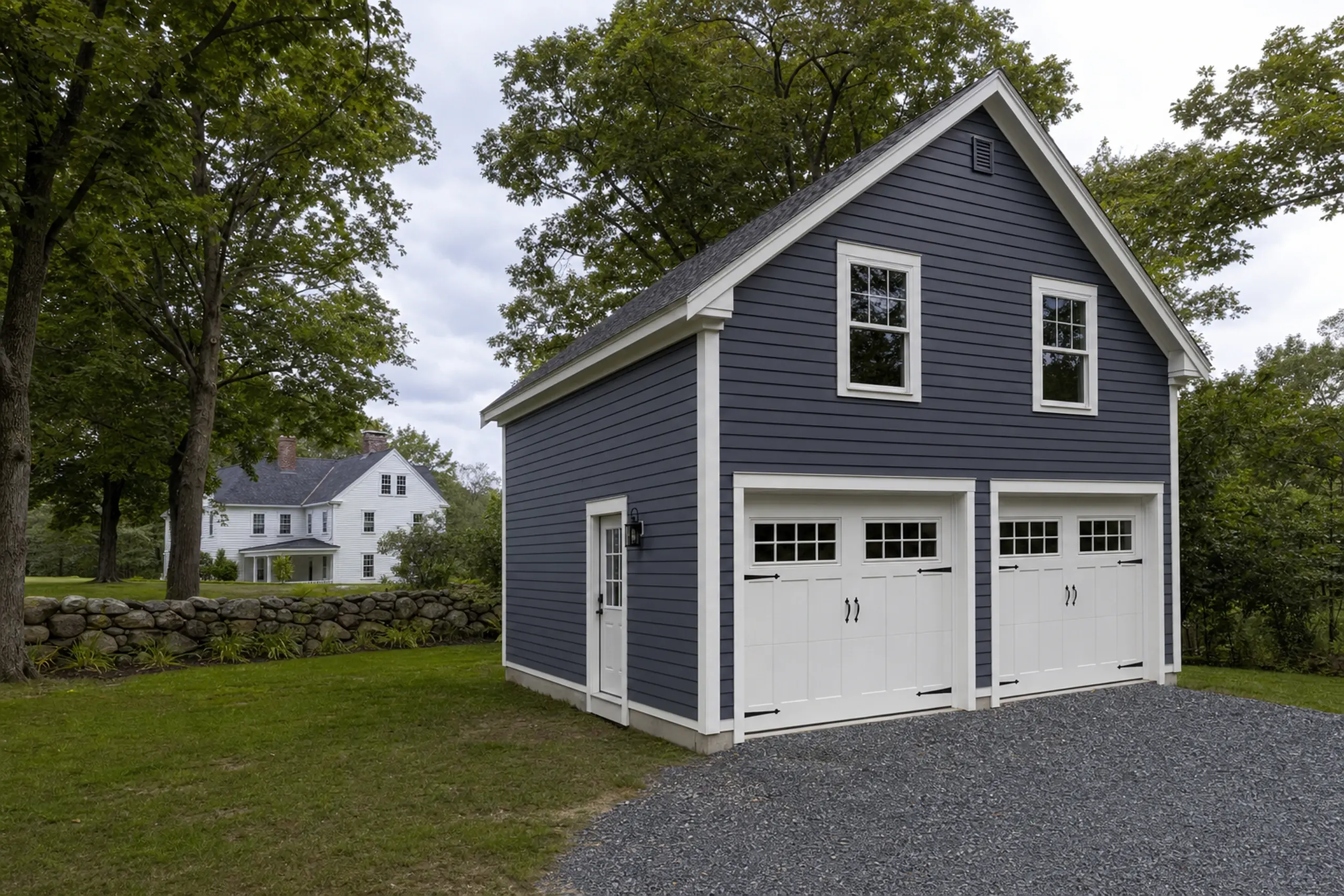 Blue two-car garage with white trim and gravel driveway in a green yard with trees.