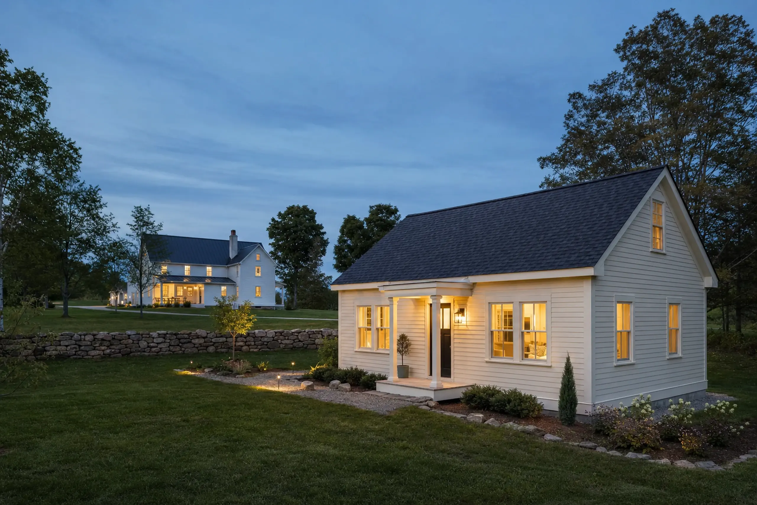 Two white houses with lit windows in a grassy, tree-lined yard at dusk.