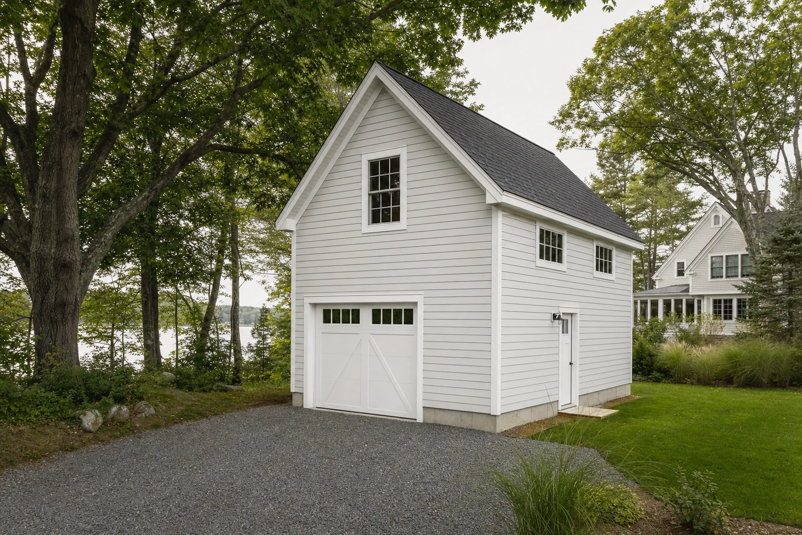 White two-story garage with black roof, surrounded by trees and grass near a gravel driveway.