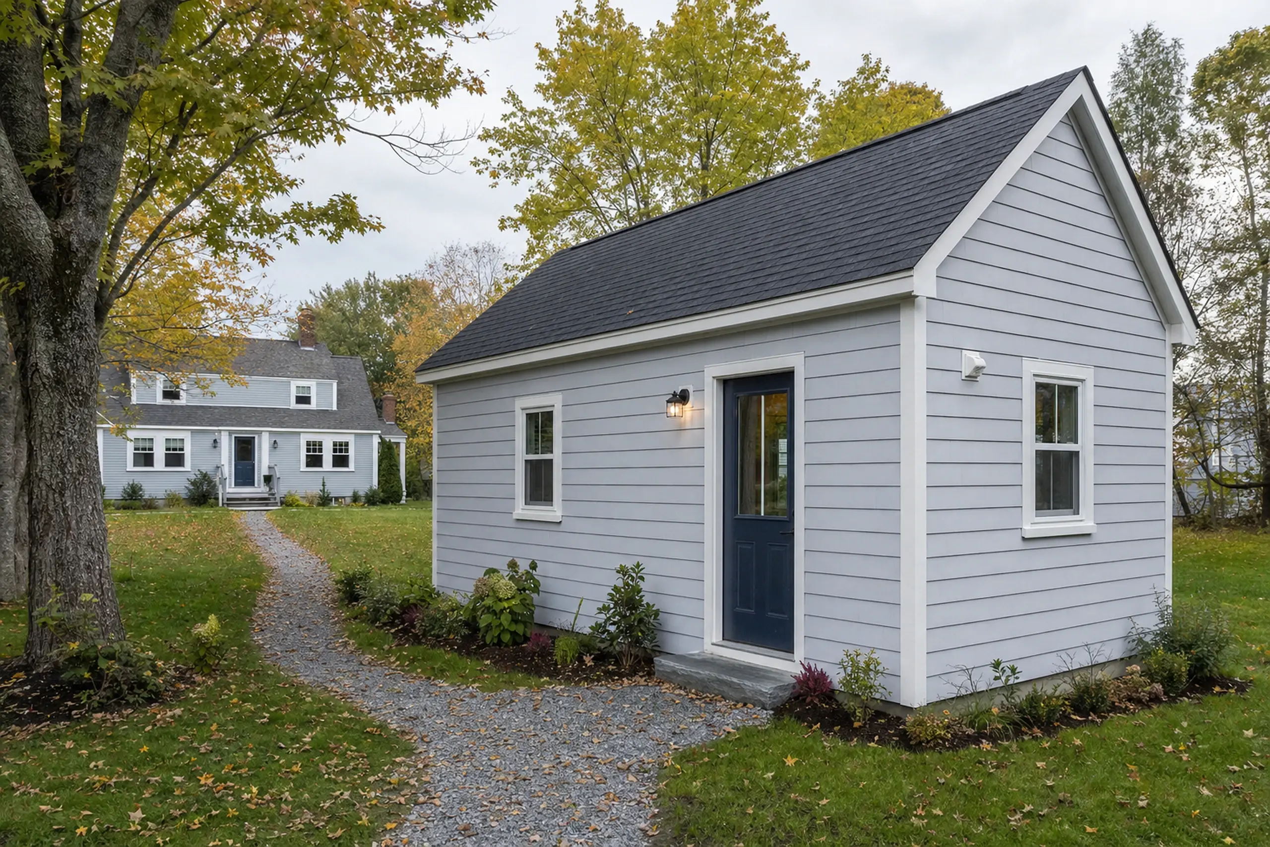 Small gray building with white trim and blue door in a grassy yard with trees and a path.