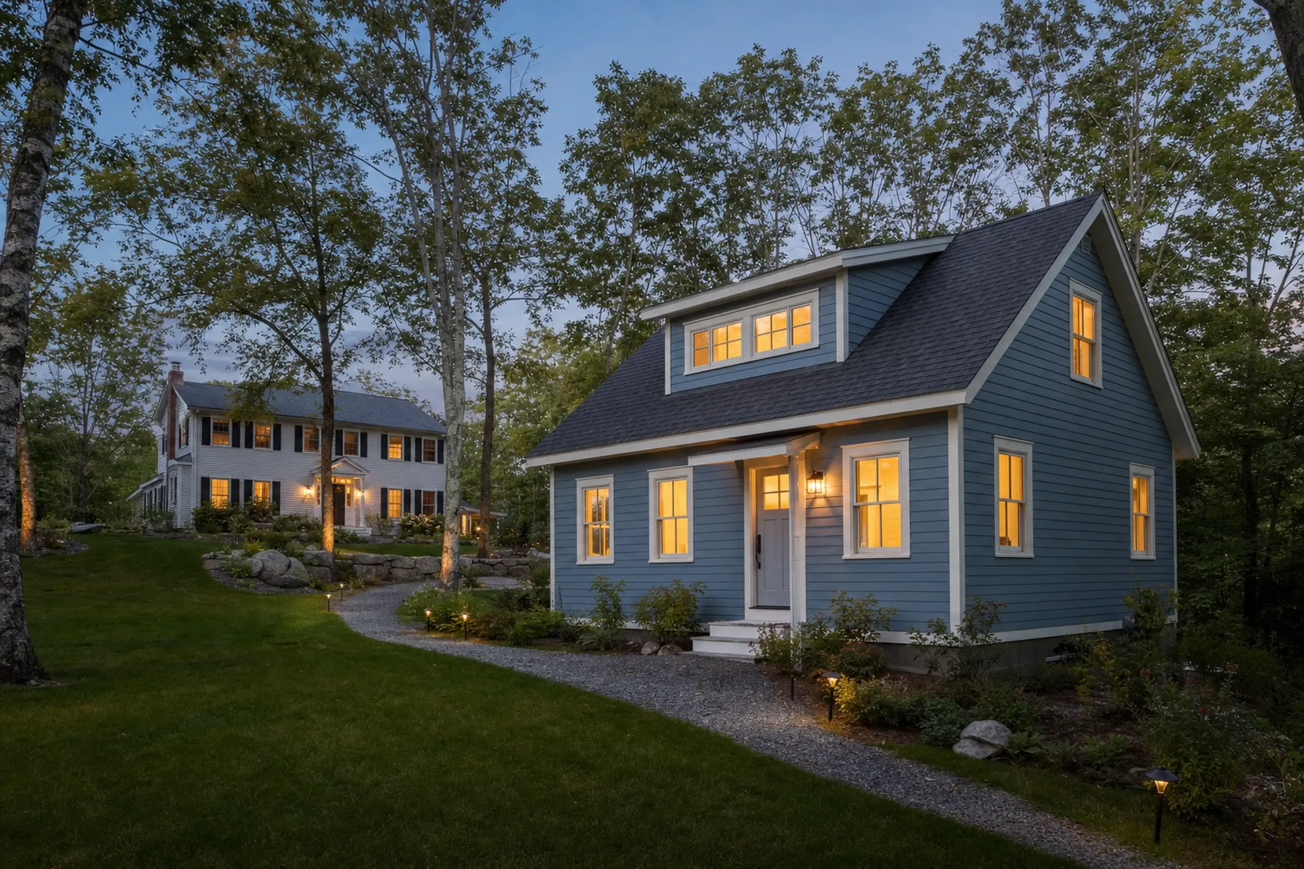 Two houses at dusk with blue and white siding, lit windows, trees, and a gravel path.