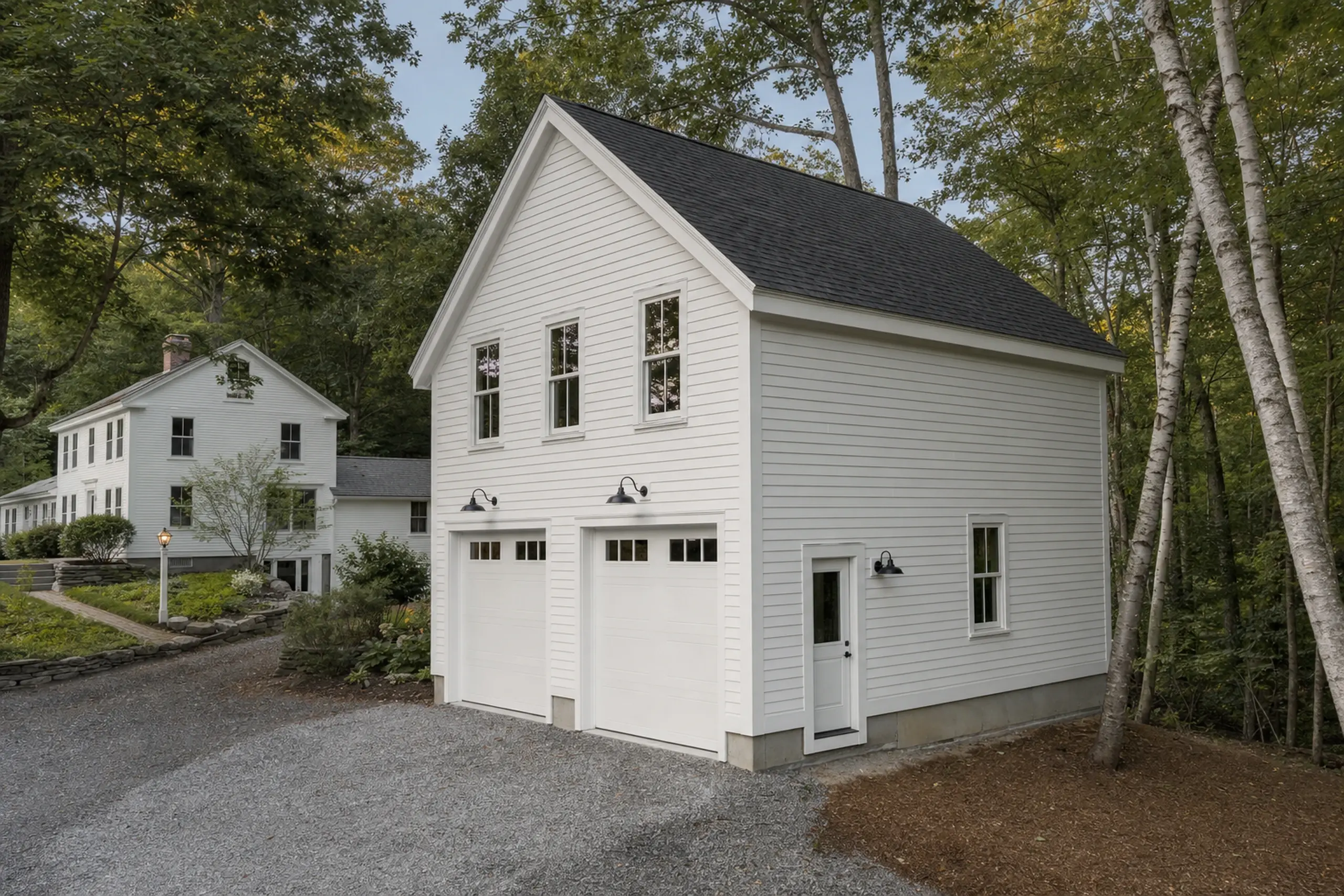 White two-story garage with black roof and two garage doors in a wooded setting.