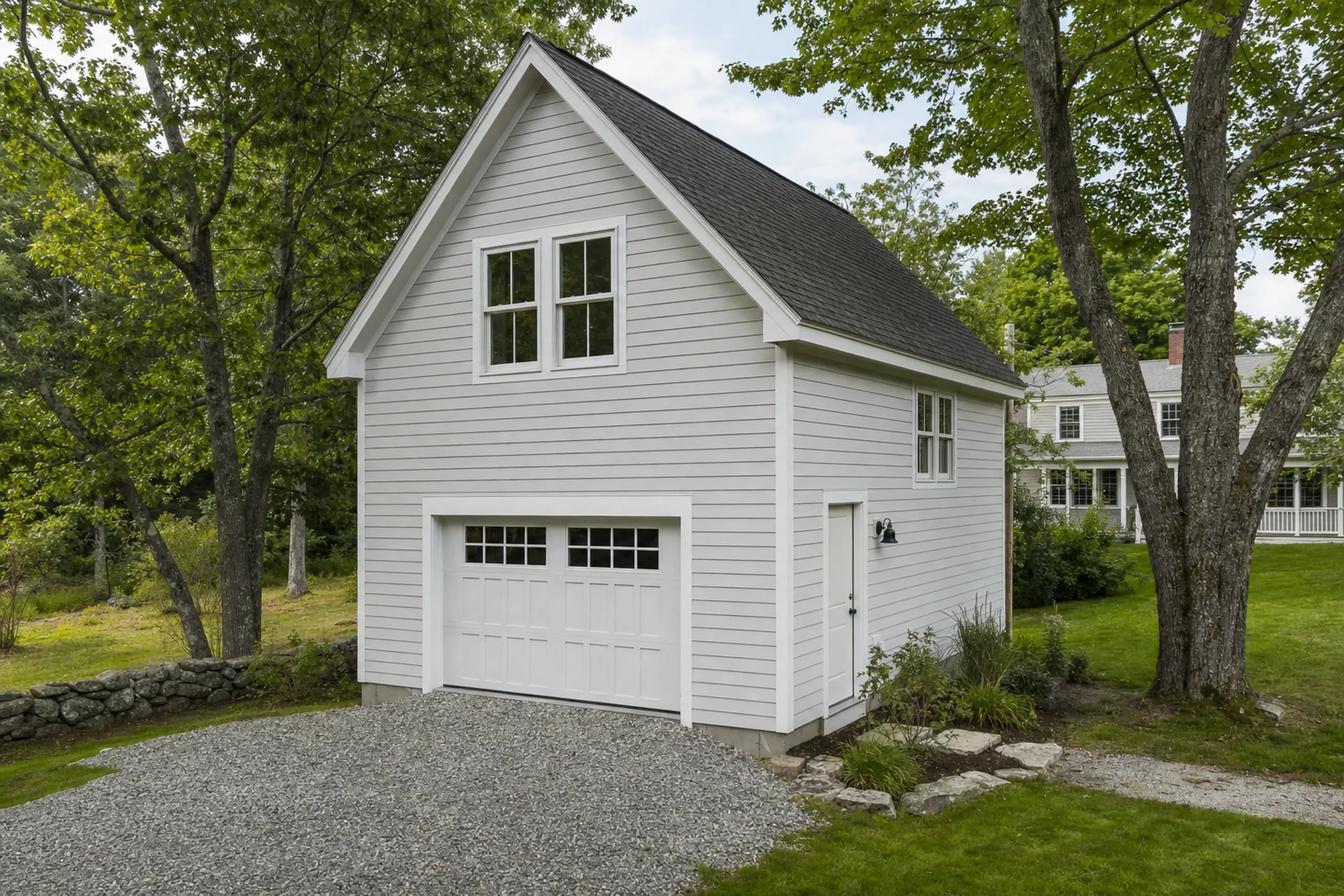 Gray two-story garage with white trim, black roof, gravel driveway, and surrounding green trees.