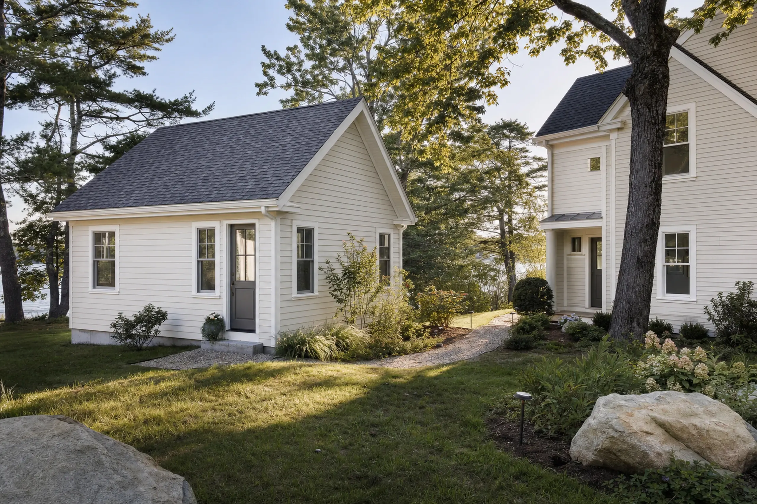 Two white buildings with gray roofs in a green yard with trees and rocks.