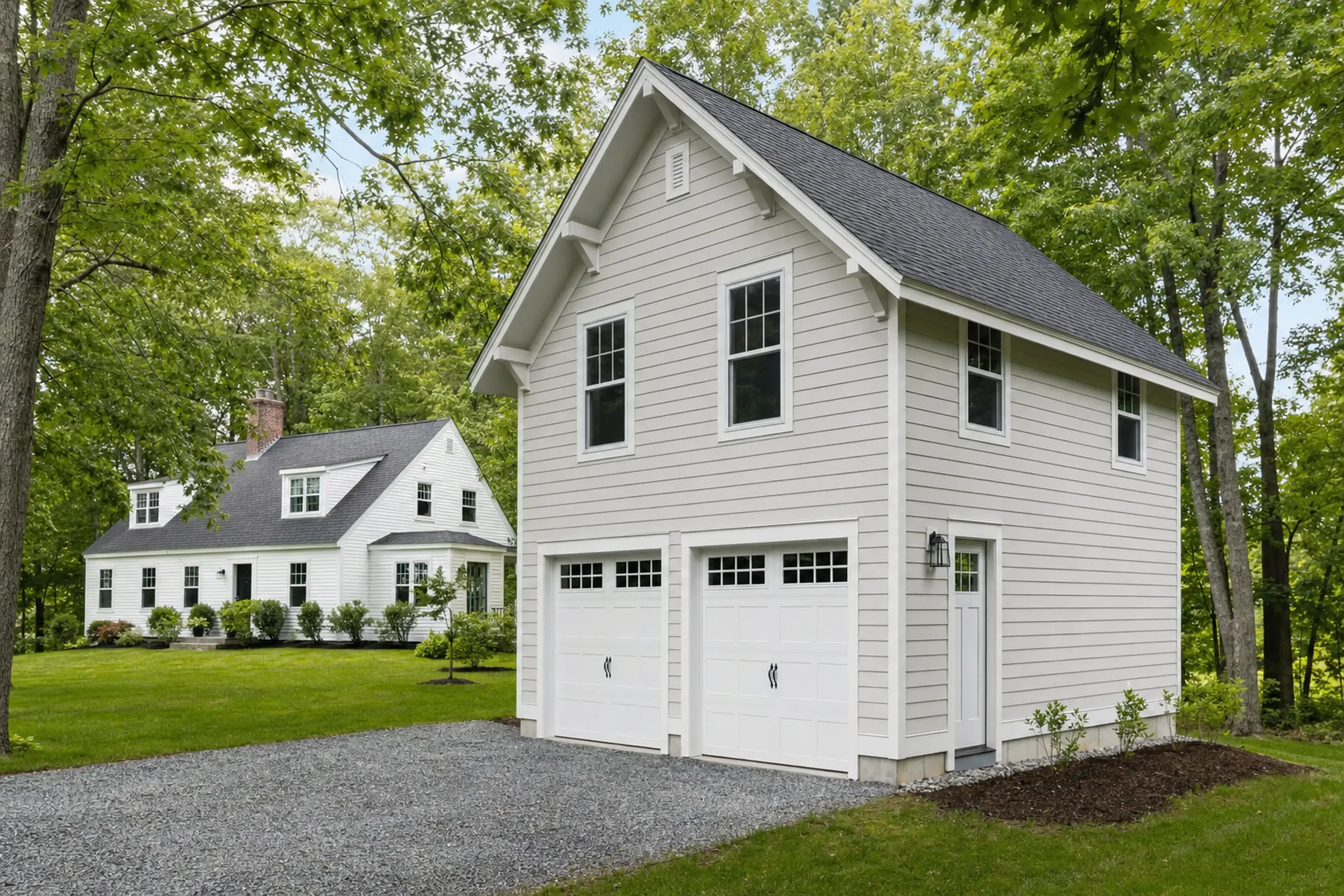 Two white buildings with black roofs in a green, tree-filled setting with gravel driveway.