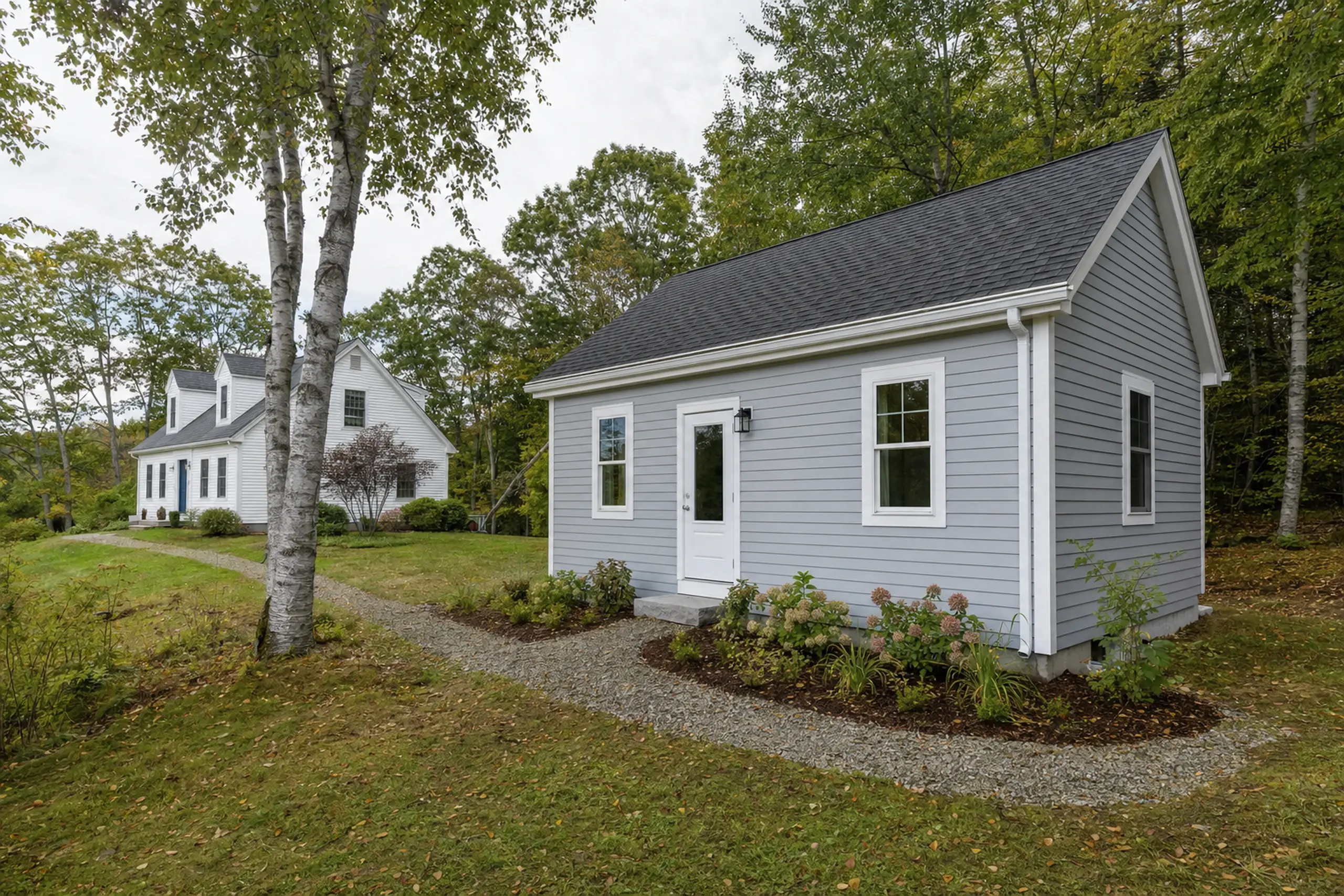 Two houses in a green yard with trees, one gray with white trim and one white in the background.