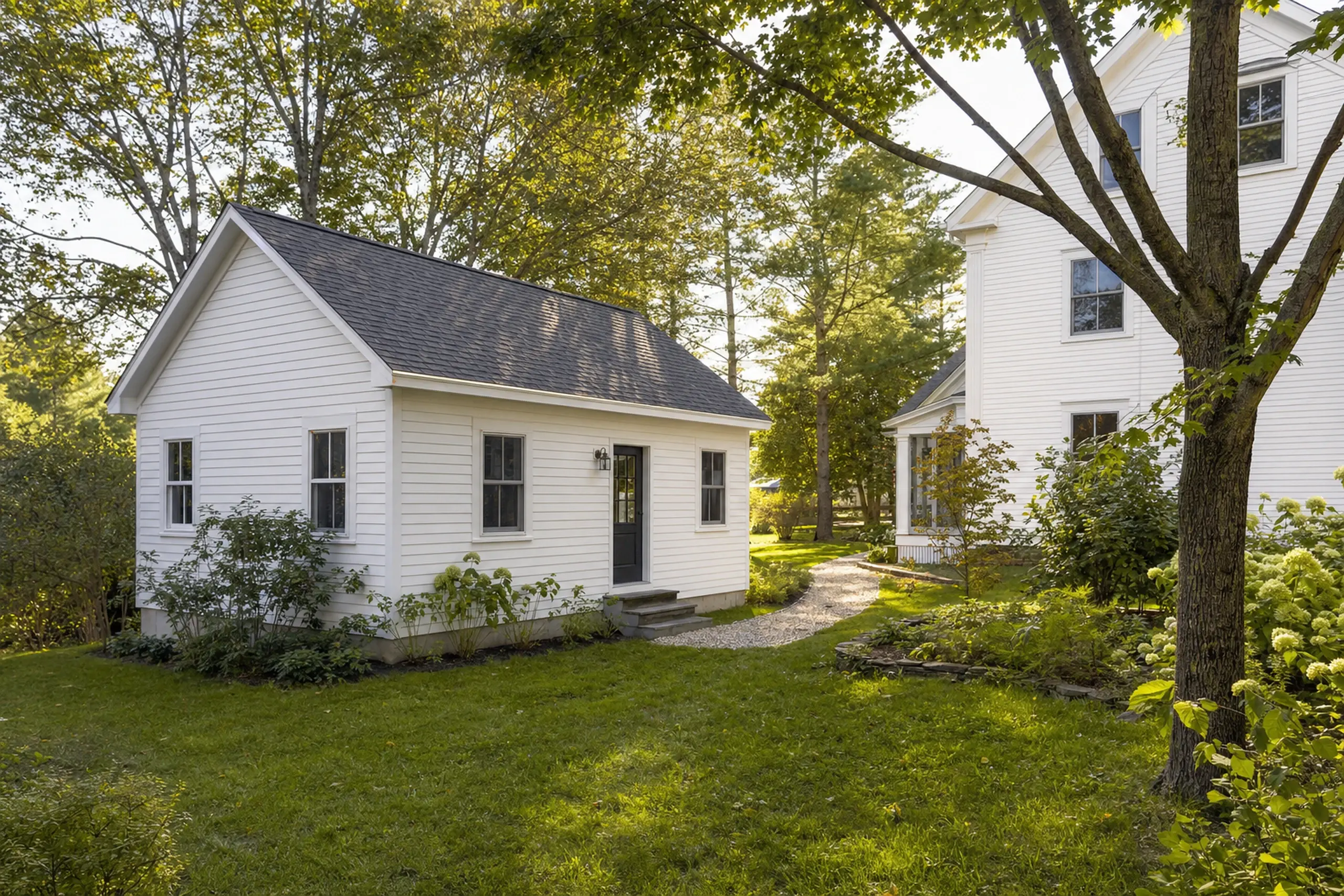 White small building with black door and windows in a green garden setting with trees.