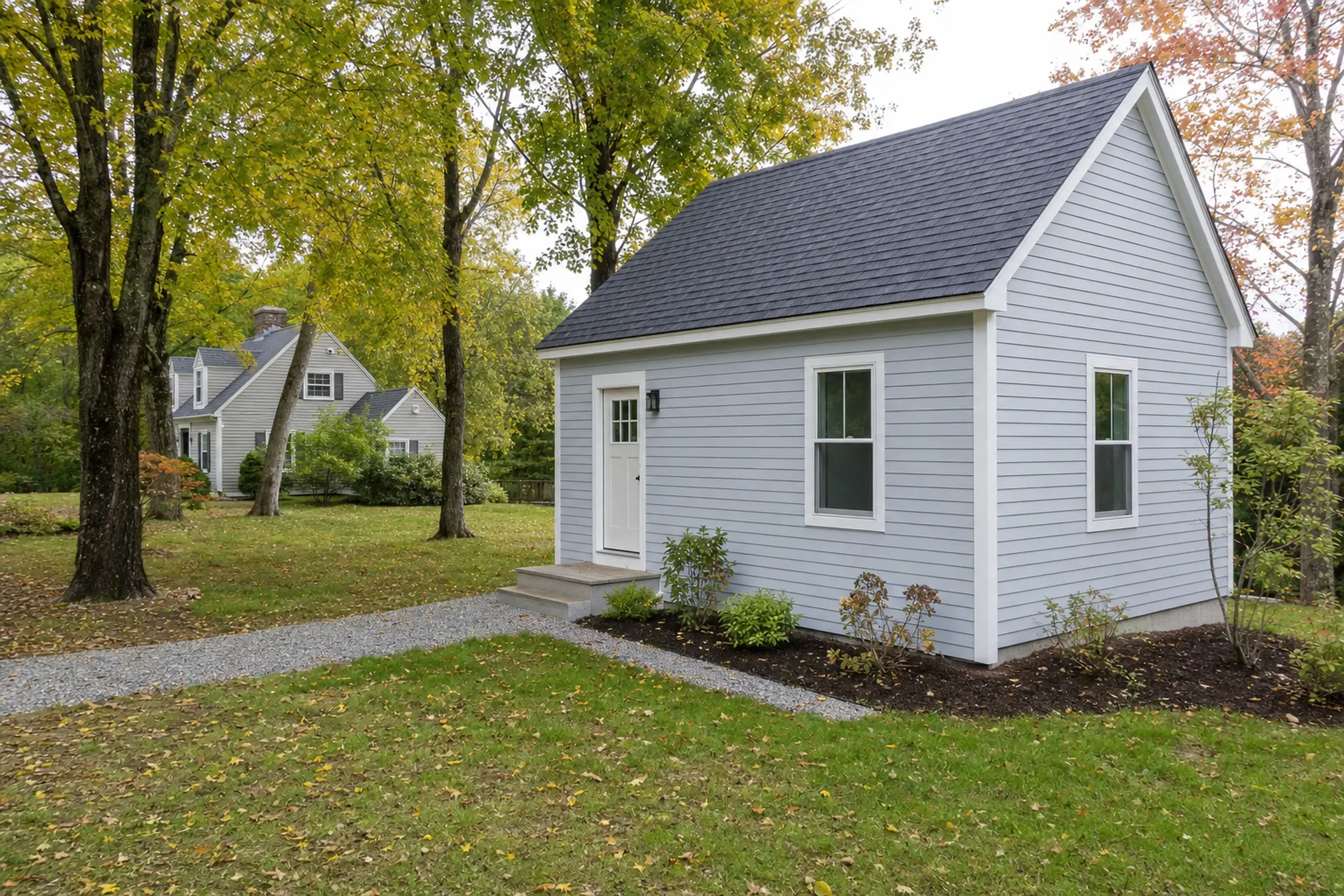 Small light blue house with white trim in a green yard with trees and a gravel path.