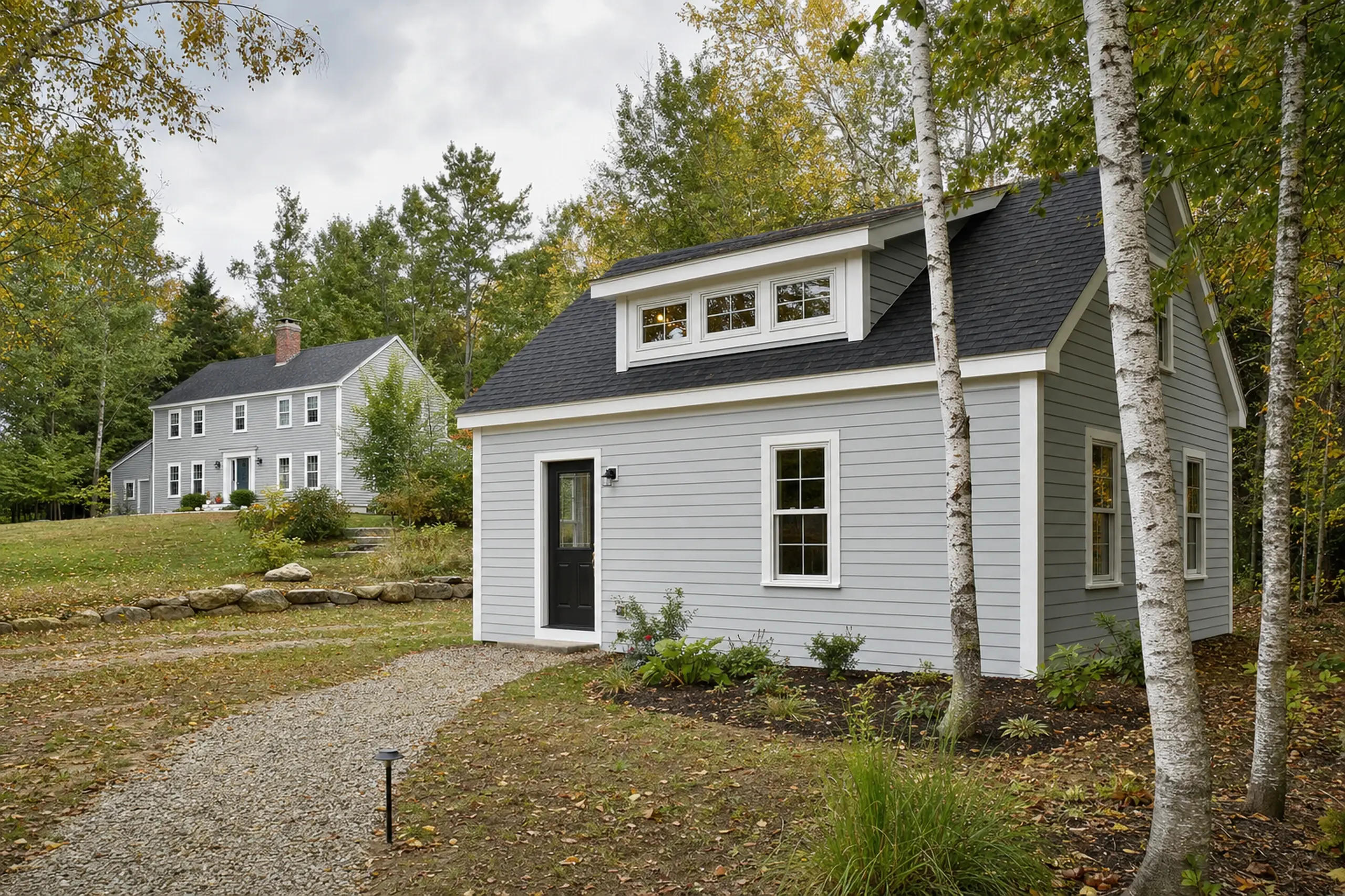 Two gray houses with white trim in a wooded setting with a gravel path and birch trees.