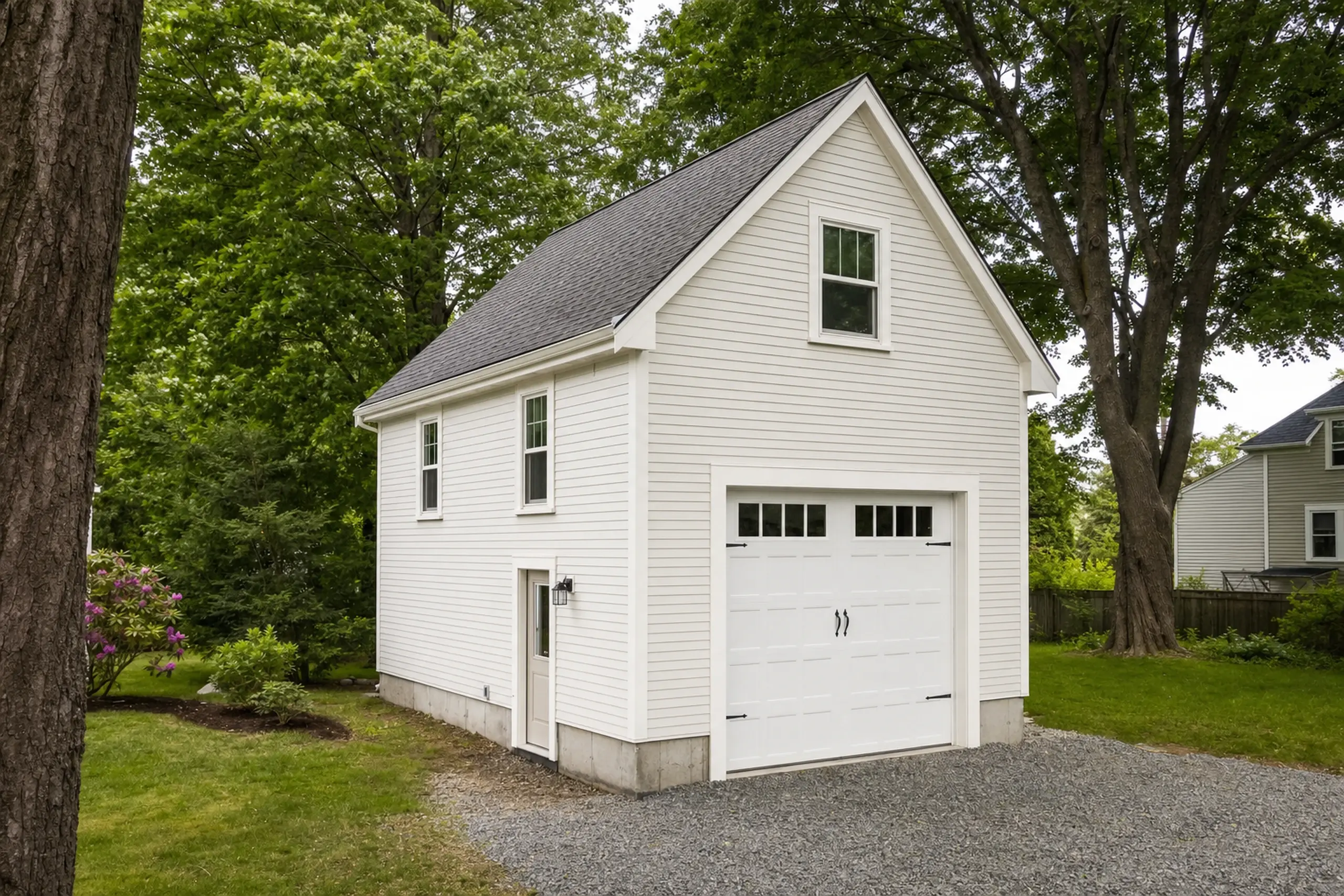 White two-story garage with gray roof, surrounded by green trees and grass.